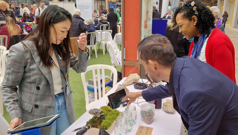 Image of one researcher and two visitors at the Flourishing & Wellbeing stand duirng the BRC's Open Day 2024. The man is taking a photograph of the moss.